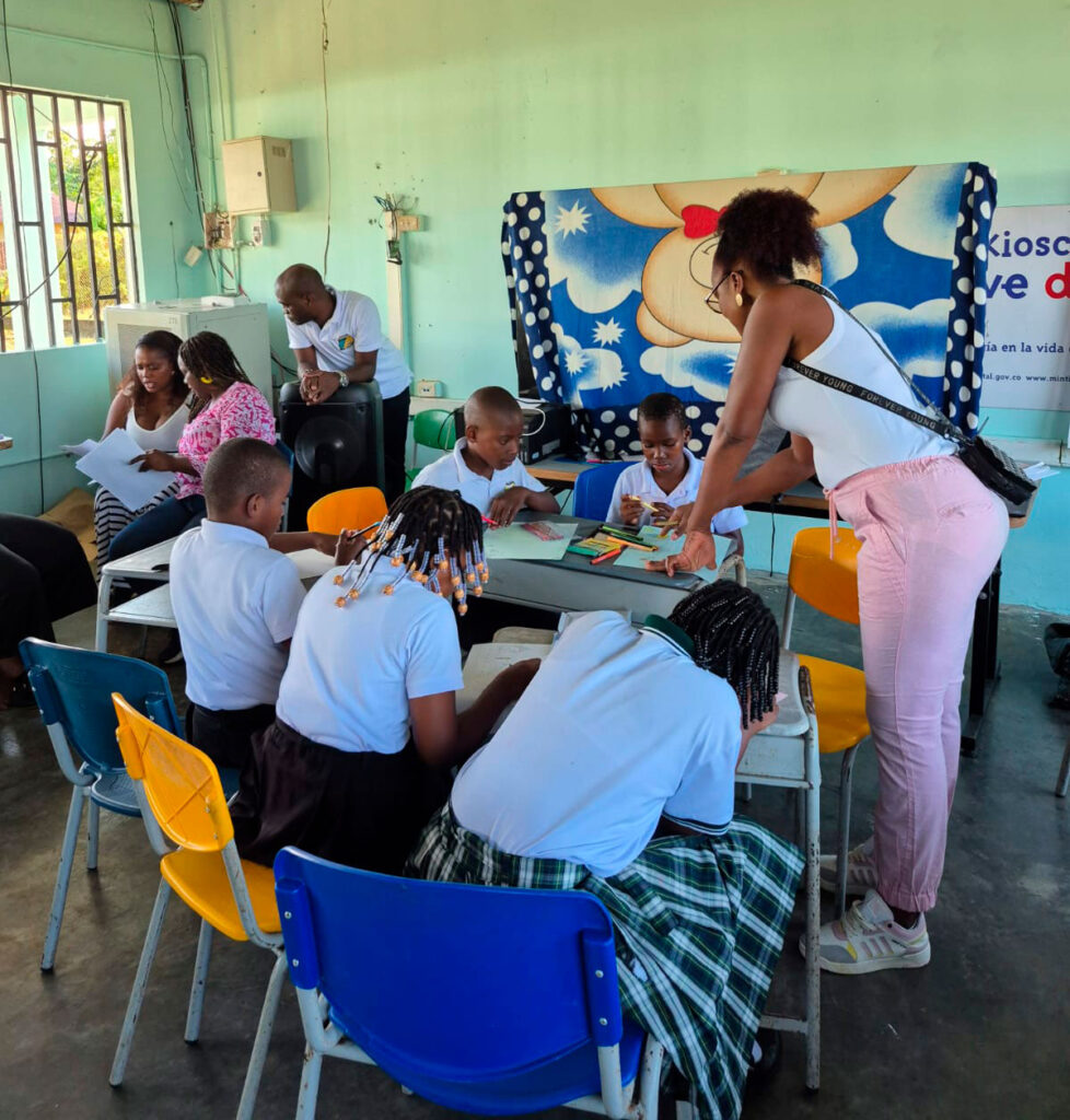 Grupo de niños y niñas de primaria trabajando en mesas circulares durante un taller creativo, acompañados por docentes en un aula con techo de lámina.